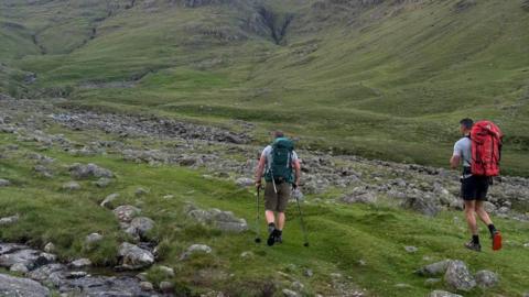 Two men walking along a flat expanse of grass with lots of rocks. They are both wearing t-shirts and shorts and the man on the right has a red walking backpack and the one on the left has a green and is using two walking poles. The ground is steep all around them.