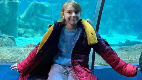 A young girl sits on the edge of a fish tank. She is blonde and wearing a purple coat, blue trousers and a blue top. She is smiling.