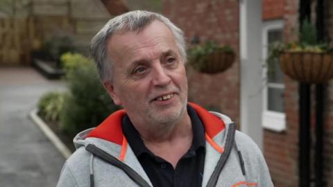 John Frosdick, who has white hair and a neat beard, can be seen wearing a grey hooded jacked over a black polo shirt as he stands outside his home. 