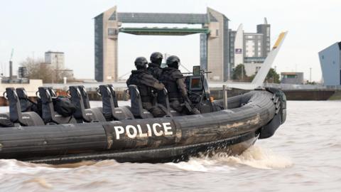 A black police rigid‑hulled inflatable boat moves quickly along a wide, brown river. Three officers in black tactical gear and helmets sit near the front. The word “POLICE” is printed in bold white letters along the side. In the background, large modern buildings and the distinctive green‑and‑white structure of Hull’s tidal barrier rise above the riverbank under a pale, overcast sky.
