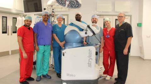 Andrew Port is a man with short white hair and glasses and is wearing dark scrubs. Next to him there are four woman and two men wearing scrubs of different colours. They are stood in a surgical room with the white and blue Mako robotic arm.