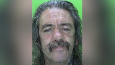 A mugshot of Jason Dyer, a 54-year-old White man with combed-back, greying long hair and a long, untidy moustache. He has thick greying eyebrows and a large nose. He appears to be frowning, his eyes are unfocused and looking downwards. Part of a tattoo appears can be seen on the left side of his neck but it is not clear what the tattoo is. The mugshot has a bright green background.