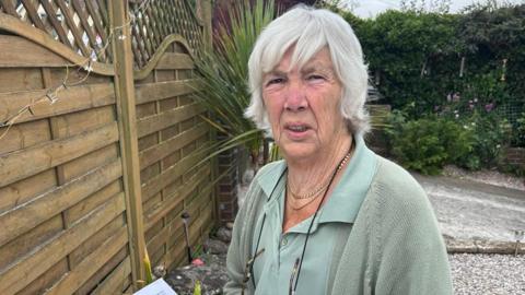 Jennie Warren looks cross while standing next to her garden fence. She is wearing a green shirt and green cardigan.