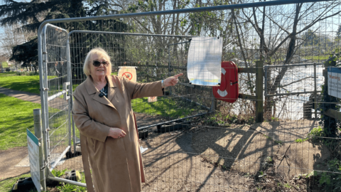 Blonde woman wearing sunglasses and brown coat next to fence and pointing at notice on it