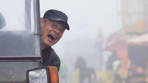 Martin Clunes laughs, mouth wide, towards the camera as he leans out the side of a utility vehicle. It's a wet, rainy day. In the background in the mist is a colourful traditional funfair. Clunes is wearing a black baseball cap and a black coat.