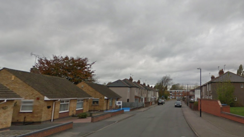 Houses and bungalows line the road with some cars parked on either side of the street.