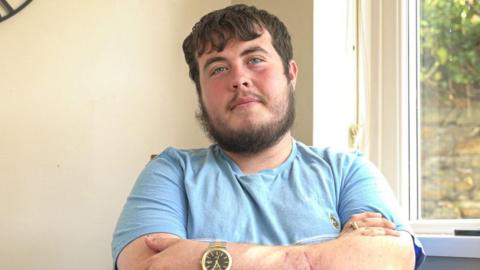 Will, who has short brown hair and wears a blue t shirt, looks at the camera with his arms folded and a neutral expression