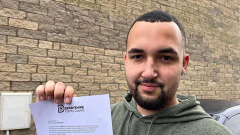 A close-up photograph of Aston Grisdale smiling and holding a confirmation from Derbyshire County Council regarding his claim for compensation. He is wearing a hoodie.