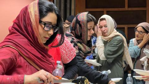 A group of women stand at a food-serving table, with one person in the foreground wearing a red outfit and serving food onto a plate. Others behind hold plates while waiting in line, with various trays and dishes laid out on the table.