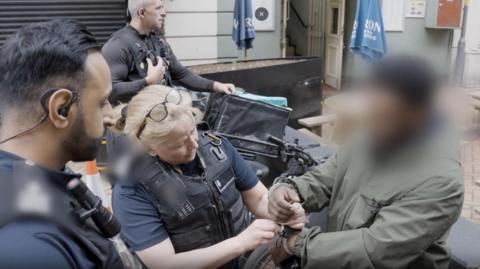 Three immigration officers putting handcuffs on a man. He has dark skin and is wearing a green jacket. There is an e-bike behind him. His face has been blurred so we do not know who he is. 
