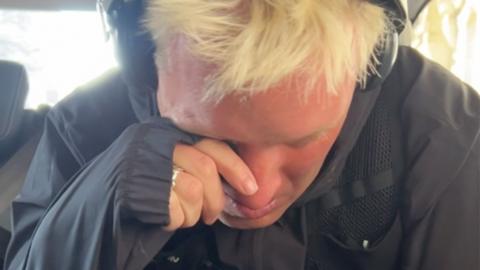 Jamie Laing, wearing headphones and sports gear, wiping tears off his face as he sits in a van