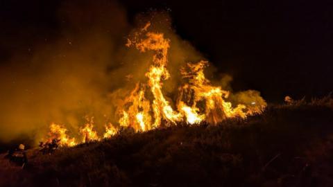 An image of tall, bright orange and black flames coming off the moorland. It is dark and the bright flames contrast against the night sky. There is also a lot of smoke coming from the area. There is a firefighter to the left of the image.