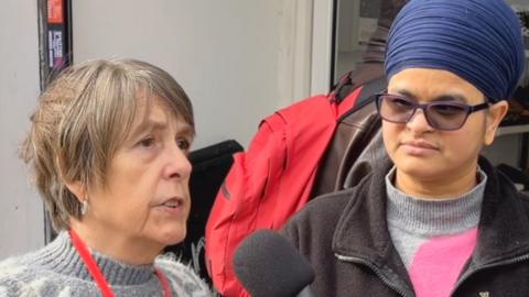 Woman on left wearing a grey jumper, has brown hair and a red lanyard talking into a microphone. Woman on right wearing a hijab, glasses, grey jumper and black cardigan listens in.