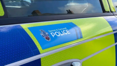 The side of a police car, blue and yellow, with the Devon & Cornwall Police sign and emblem on the left of the writing.