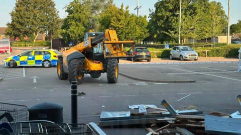 A yellow JCB telescopic handler stands in front of the damaged bollards, wall and windows of the Tesco car park. A police car is parked behind blue and white police tape.