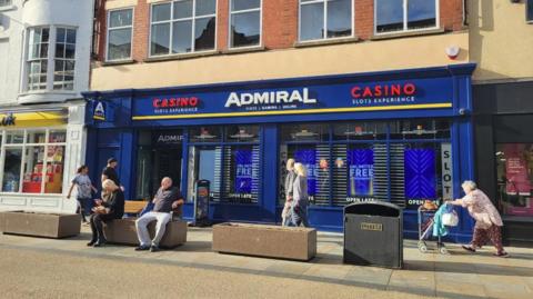 A gaming centre shop front which is bright blue with red and white lettering. There are seats outside with a man and woman sitting down. A woman pushes a mobility scooter behind them. 