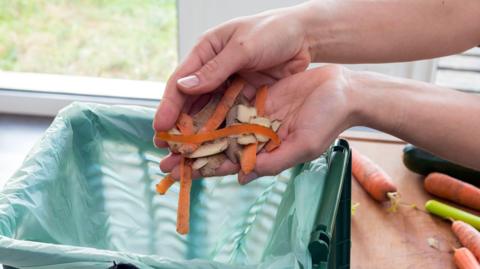 A close-up of a person's hands holding carrot and potato peelings above a green food waste bag. More carrots can be seen on a chopping board.