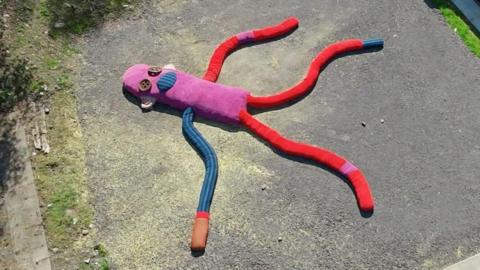 A sculpture of a giant sock puppet lying in a car park. It has a pink body, one blue and one red arm and two red legs. It has two giant button shapes for eyes.