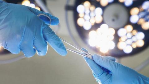 Two hands in surgical gloves with a pair of scissors being handed over in an operating theatre setting.