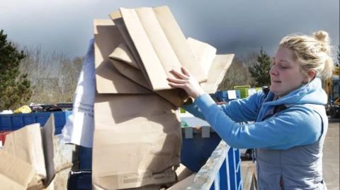 A woman with blonde hair wearing a blue sleeveless jacket over a blue hooded top puts cardboard into a blue skip