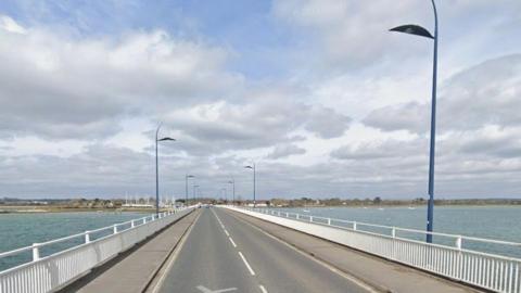 Road bridge with white railings and dark blue lights over water.