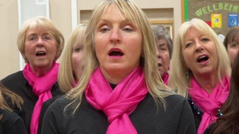 Annabelle Underwood - a blonde woman wearing black clothing with a pink scarf. Her mouth is open as she sngs. She has other choir members around her all singing too.