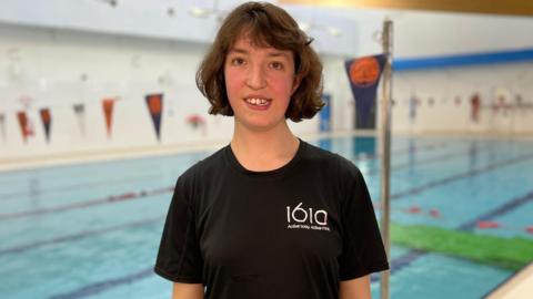 Emily Gold. She has dark short hair and is pictured standing in front of a leisure centre swimming pool. She is wearing a black t-shirt with 1610 on the chest. She is looking directly at the camera and smiling.