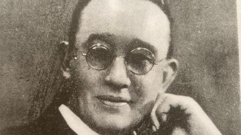 An old black-and-white portrait of a man in a priest's white collar posing with a hand on his chin. He is smiling and wearing round glasses and his dark hair is swept back from his face.