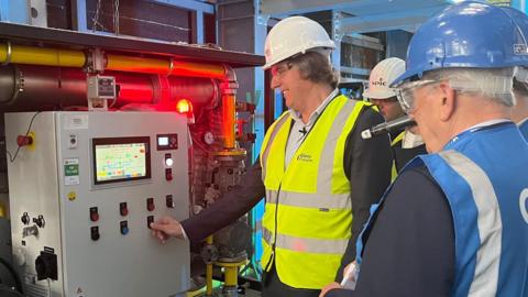 Steve Rotheram is wearing a black suit with a blue shirt and a high-visibility yellow jacket and white hard hat. He is pressing the button on a large grey metal box control system. He is stood alongside two people wearing blue high-visibility jackets and blue hard hats.