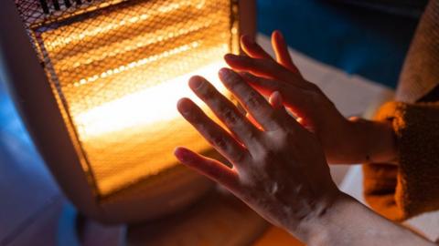 A close-up of hands being warmed at an electric heater. The heater is burning dep red, the person appears to be female and wearing a house coat. of indeterminate light colour.