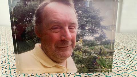 A family photograph is on display in the family home. Ian Massey is smiling at the camera and is wearing a yellow top. Trees are in the background.