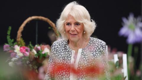 Queen Camilla who is standing amongst flowers and bouquets of flowers at the Sandringham Flower Show. She is smiling but not looking directly at the camera. She is wearing a black and white floral dress.