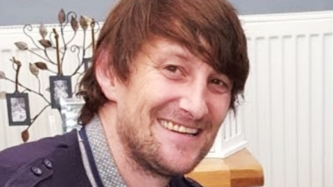 Head and shoulders shot of Andrew Hamilton, a white man with brown hair, swept slightly to the side in a long fringe and neck length. He has a lightly-grown beard. He is wearing what looks like a polo shirt with a blue jacket over it. He is smiling at the camera. Behind him is a white radiator, a wooden side table with ornaments and photos on it. 