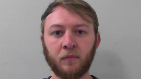 A man with a pink face, flat light brown hair and a beard, stares at the camera in a police mug shot.