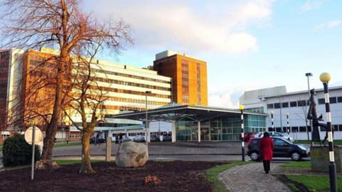 The front entrance of Altnagelvin hospital in Londonderry. A woman in a red coat can be seen walking past a statue while a number of parked cars sit outside the hospital entrance. It is dry and sunny, two trees are in the foreground, a number of hospital buildings in the background.