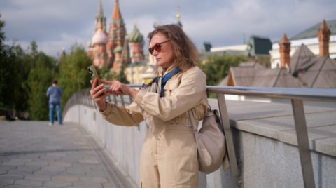 A woman in a beige mac and sunglasses uses her phone in front of the Kremlin.