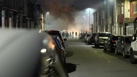 A street in Harehills, Leeds, with cars parked on both sides of the road. In the distance, people are stood around a burning bus. 