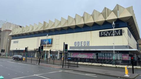 The south wall of the Kingwest building in Brighton. It is a white building with large white triangles on the roof. The word PRYZM and ODEON are written on the front. There is a horizontal blue stripe around the top quarter of the building