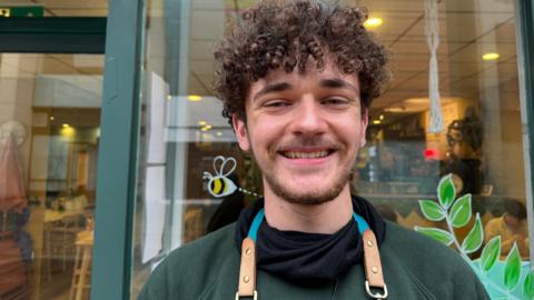 Brandon Elliott, supervisor at The Ugly Duckling Pottery Painting, smiling in front of his shop, The windows behind him are decorated with bees and tree branches. There are people inside the shop.