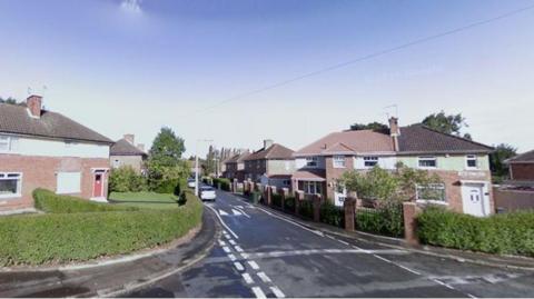 A quiet suburban street with semi-detached brick houses on both sides. The houses have tiled roofs, chimneys, and small front gardens bordered by hedges. The road is a two-way street with white dashed and solid markings, curving slightly to the left. A single white car is visible further down the street, and there are trees and streetlights in the background under a clear blue sky.