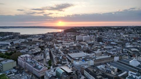A birds eye view St Helier Jersey. There is are many buildings. The sun is setting and there are grey clouds in the sky. The sea is blue. 
