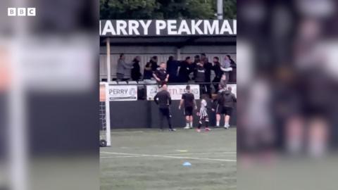 People standing in a football stand wearing black clothing. The stand is next to the football pitch