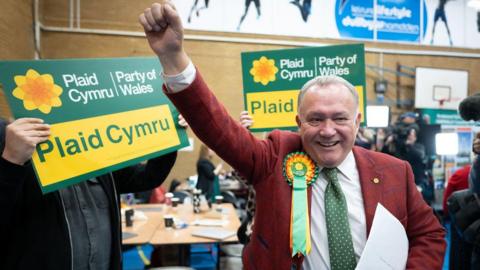 Plaid Cymru candidate Lindsay Whittle holds his fist in the air in celebration next to a Plaid Cymru sign