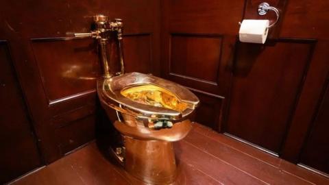 The golden toilet sitting in a corner of a room panelled with dark wood.