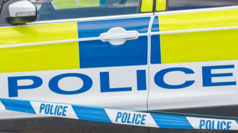 A close-up of the side of a police vehicle and the word police, with police tape in front of the car. You can see the left wing mirror and the left doors.