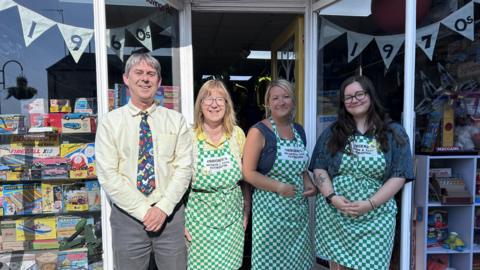 One man and three women stand outside a toy shop. All are smiling. The man is wearing a shirt and brightly-coloured tie and the women are wearing green and white checked aprons. The shop window is decorated by bunting, reading "1960s" and "1970s". Various vintage toys and games are displayed.