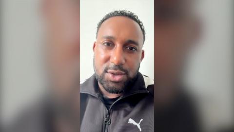 A man with short black hair, moustache and beard talks to the camera. He is wearing a zipped up sports jacket.