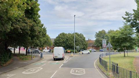 A Google Maps screen shot of Arundel Street, at the junction with Holbrook Road. The road leads to a roundabout with trees in the middle and hedging on its circumference. There are tress on each side of the road and a white van driving towards the roundabout