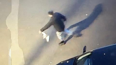 A slightly blurred CCTV image showing a man wearing black trainers, grey joggers and a black coat holding a pigeon by one wing.