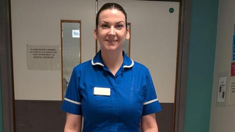 A smiling woman in a blue nurse's uniform with white trim standing in front of double doors in a hospital (Sheffield's Royal Hallamshire Hospital)
